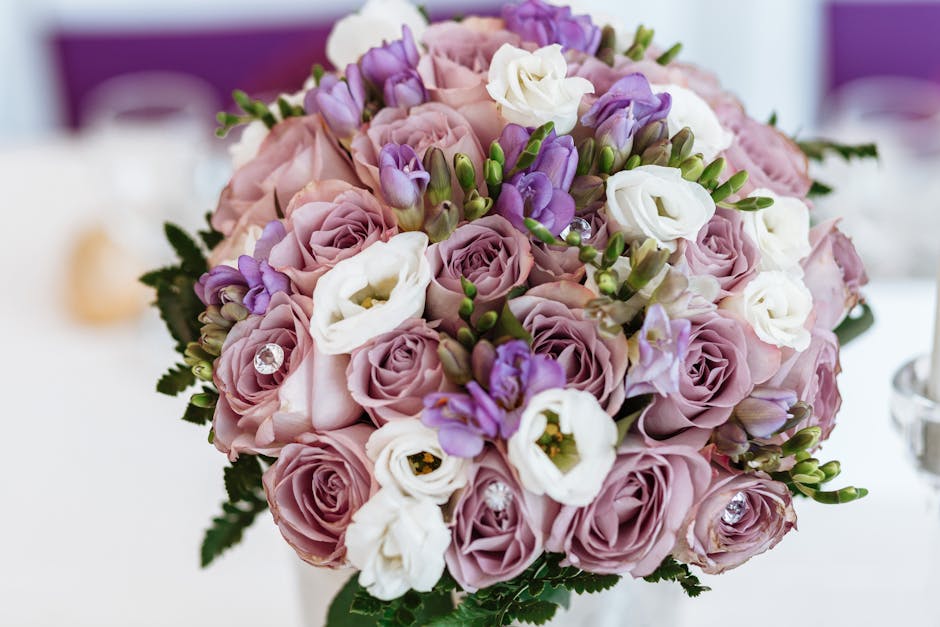 A close-up view of a floral bouquet arranged with pastel pink roses, white eustoma, and purple lisianthus, accented with green foliage. The flowers are fresh with soft, velvety petals and sturdy stems