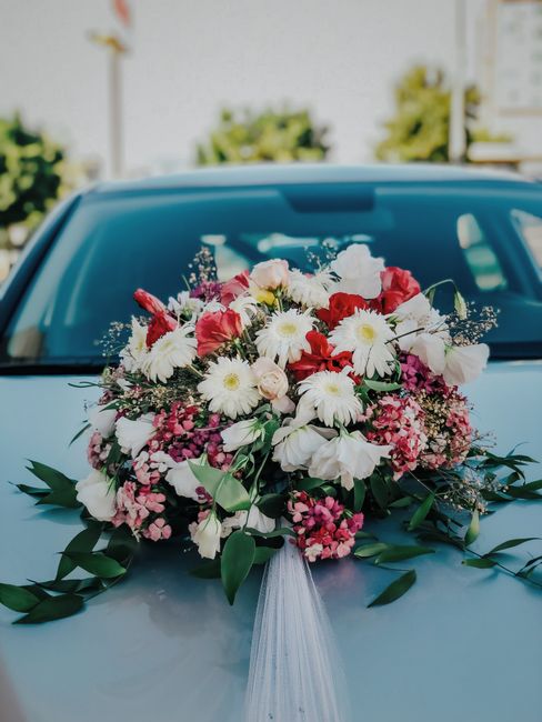 A large, vibrant floral arrangement displayed on the hood of a light blue vehicle, featuring a variety of fresh flowers including white daisies, pink and red roses, purple hydrangeas, and white lilies