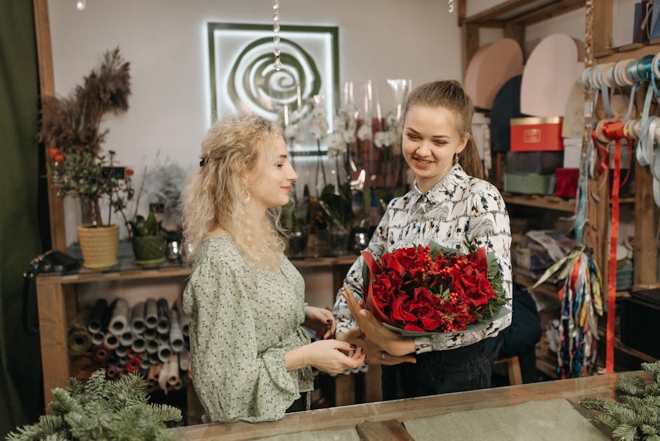 A young female florist with light brown hair tied back, wearing a white shirt with black floral patterns, presenting a vibrant bouquet of deep red roses, red alstroemeria, and green foliage wrapped in