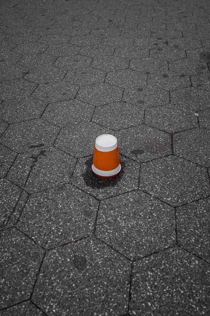 A small orange and white safety cone placed on a textured hexagonal paved surface outdoors. The cone appears to be made of plastic with a smooth finish, and it is positioned centrally in the image. Th
