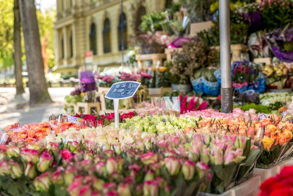A vibrant outdoor flower stall featuring a wide display of fresh flowers arranged in bouquets and bunches, predominantly roses in shades of pink, cream, and peach, with some orange blooms. The flowers