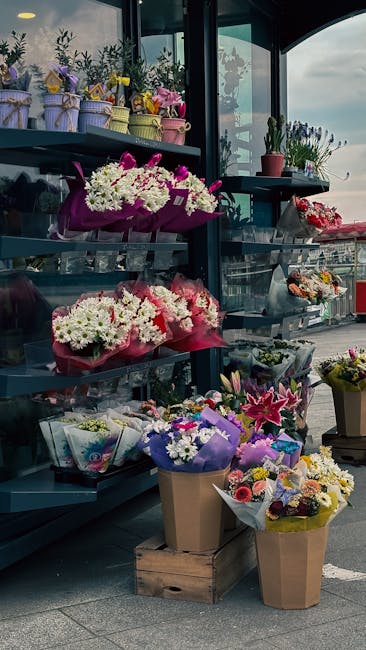 A display of fresh floral arrangements and bouquets arranged on dark-tiered shelves outside a flower shop, featuring a variety of flowers including white chrysanthemums, pink lilies, purple irises, an