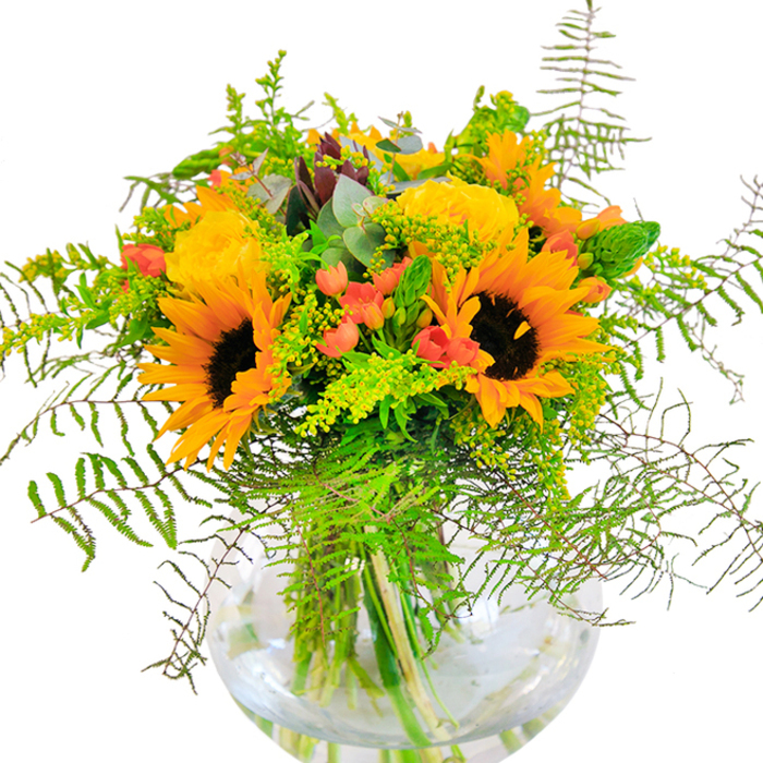 Overflowing with brightness, this exquisite floral arrangement is a tribute to the cheerful days of Wembley, capturing both nature's artistry and the skill of local florists. Dominating the bouquet, broad-faced sunflowers radiate golden warmth, their bold petals framing rich chocolate-brown centers. Interlaced with them are exquisite yellow roses, their petals tightly layered into luminous, rosette shapes that evoke beauty and celebration. Glossy red Hypericum berries dot the design, adding a touch of playfulness reminiscent of the vibrant stalls at Wembley Market. A bounty of greenery - from cascading ferns to airy sprigs of goldenrod flecked with delicate yellow flowers - weaves through the blooms, introducing depth and movement much like the leafy tranquility found along Brent River Park. The glass vase, crystal-clear and perfectly round, cradles each stem in cool water, magnifying the freshness and emphasizing the architectural grace of the arrangement. Eucalyptus leaves lend subtle scents and a gentle blue-green accent, completing the rustic yet elegant feel. This bouquet is ideal for birthdays and festive gatherings in Wembley, its palette and textures bringing a joyful, sunlit radiance to any space. Every detail attests to the care and craftsmanship of Flowers Wembley, reflecting the heart and artistry of the local community.